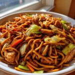 A close-up of a generous serving of homemade chicken chow mein recipe, featuring noodles, chicken pieces, carrots, and cabbage.