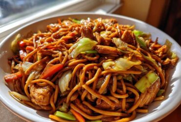 A close-up of a generous serving of homemade chicken chow mein recipe, featuring noodles, chicken pieces, carrots, and cabbage.