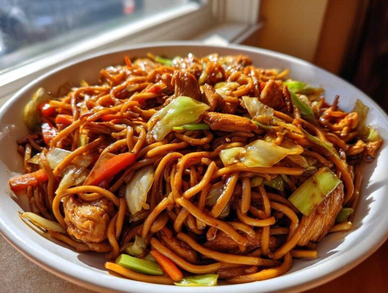 A close-up of a generous serving of homemade chicken chow mein recipe, featuring noodles, chicken pieces, carrots, and cabbage.