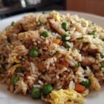 A close-up serving of amazing chicken fried rice featuring white rice, chunks of chicken, peas, carrots, and scrambled egg.