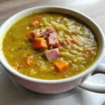A close-up of a white mug filled with thick, yellow-green pea soup, topped with diced ham and carrots.