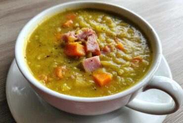 A close-up of a white mug filled with thick, yellow-green pea soup, topped with diced ham and carrots.