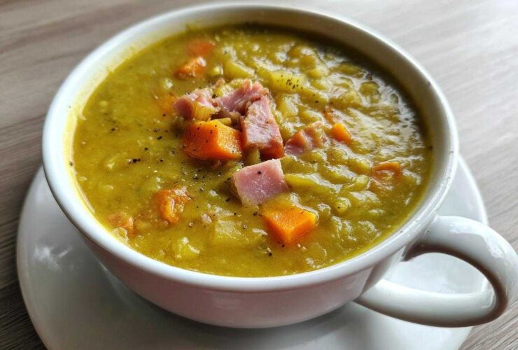 A close-up of a white mug filled with thick, yellow-green pea soup, topped with diced ham and carrots.