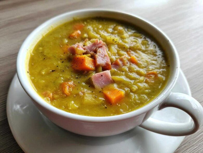A close-up of a white mug filled with thick, yellow-green pea soup, topped with diced ham and carrots.