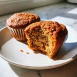 Close-up of a moist banana protein muffin with a bite taken out, revealing the texture, next to a whole muffin.