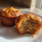Two golden-brown banana protein muffins on a white plate, one with a bite taken out showing the moist interior.