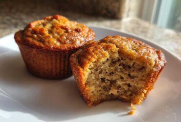 Two golden-brown banana protein muffins on a white plate, one with a bite taken out showing the moist interior.
