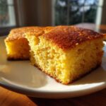 Two golden-brown squares of moist buttermilk cornbread resting on a white plate, highlighted by sunlight.