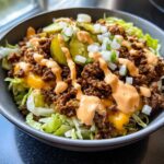 A close-up view of a hearty cheeseburger bowl featuring seasoned ground beef, melted cheese, lettuce, pickles, and a drizzle of special sauce.