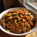 A close-up of a white bowl filled with delicious chicken chow mein recipe, featuring noodles, chicken strips, carrots, and cabbage.