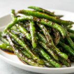 Close-up of vibrant, blistered chinese green beans with garlic sauce piled on a white plate.