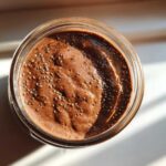 Overhead view of rich chocolate overnight oats topped with chia seeds in a glass jar, illuminated by sunlight.