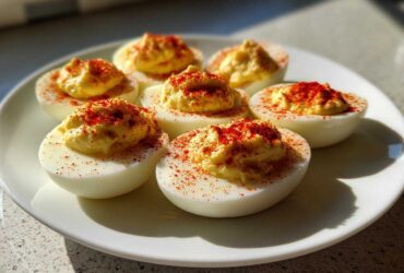 A white plate holding six creamy, perfectly piped deviled eggs, generously dusted with bright red paprika.