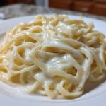 Close-up of fettuccine pasta generously coated in rich, creamy homemade alfredo sauce on a white plate.