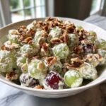 Close-up of a bowl filled with creamy grape salad, featuring green and red grapes coated in dressing and topped with chopped pecans.