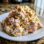 A close-up of a generous serving of creamy ham salad mixed with pickles and dressing on a white plate.