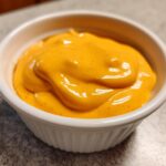 Close-up of smooth, bright orange nacho cheese sauce swirled in a small white dipping bowl.