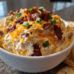 A close-up of creamy Million Dollar Dip served in a white bowl, topped generously with shredded cheddar, bacon bits, and green onions.