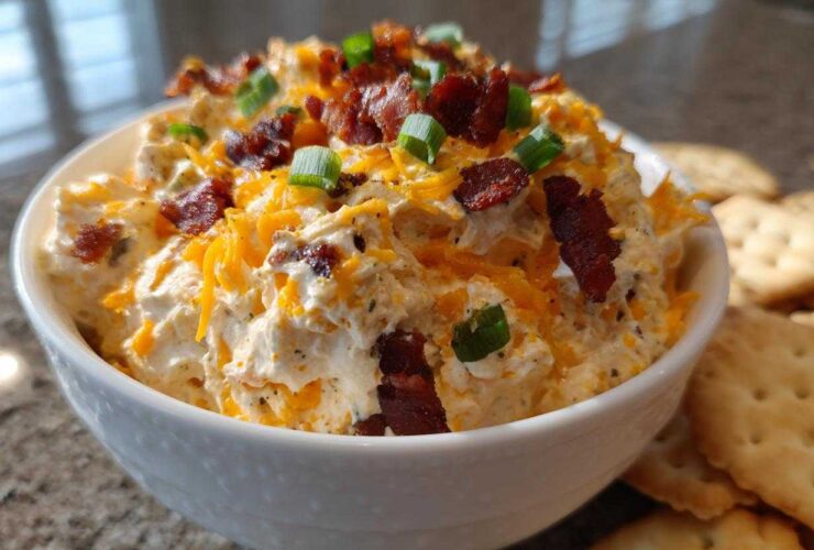 A close-up of creamy Million Dollar Dip served in a white bowl, topped generously with shredded cheddar, bacon bits, and green onions.