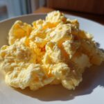 A close-up of fluffy, creamy scrambled eggs piled high on a white plate, highlighted by natural sunlight.