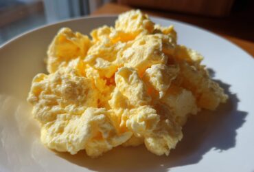 A close-up of fluffy, creamy scrambled eggs piled high on a white plate, highlighted by natural sunlight.