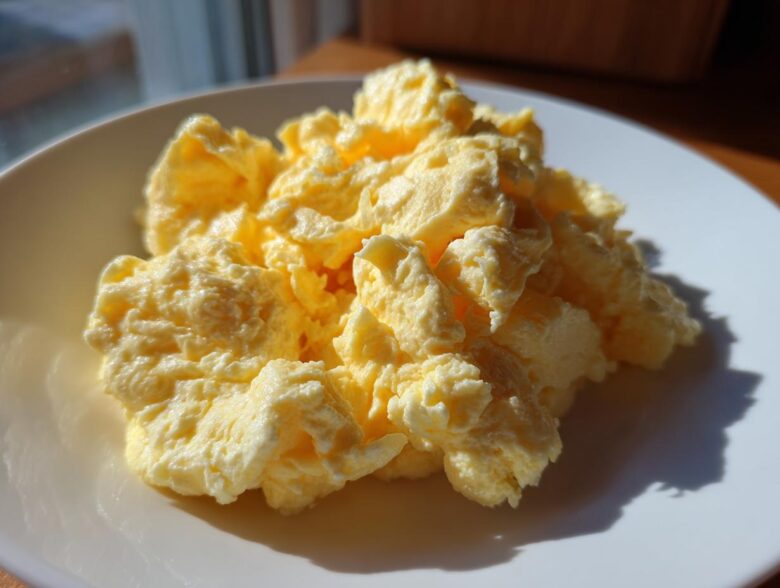A close-up of fluffy, creamy scrambled eggs piled high on a white plate, highlighted by natural sunlight.