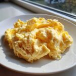 Close-up of fluffy, creamy scrambled eggs served on a white plate, highlighted by natural sunlight.