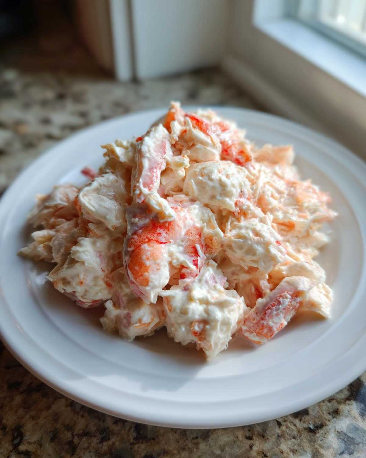 A close-up of a mound of creamy seafood salad featuring chunks of pink and white seafood mixed in a white dressing, served on a white plate.