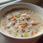 Close-up of a white bowl filled with creamy chicken and vegetable wild rice soup, featuring shredded chicken and visible wild rice grains.