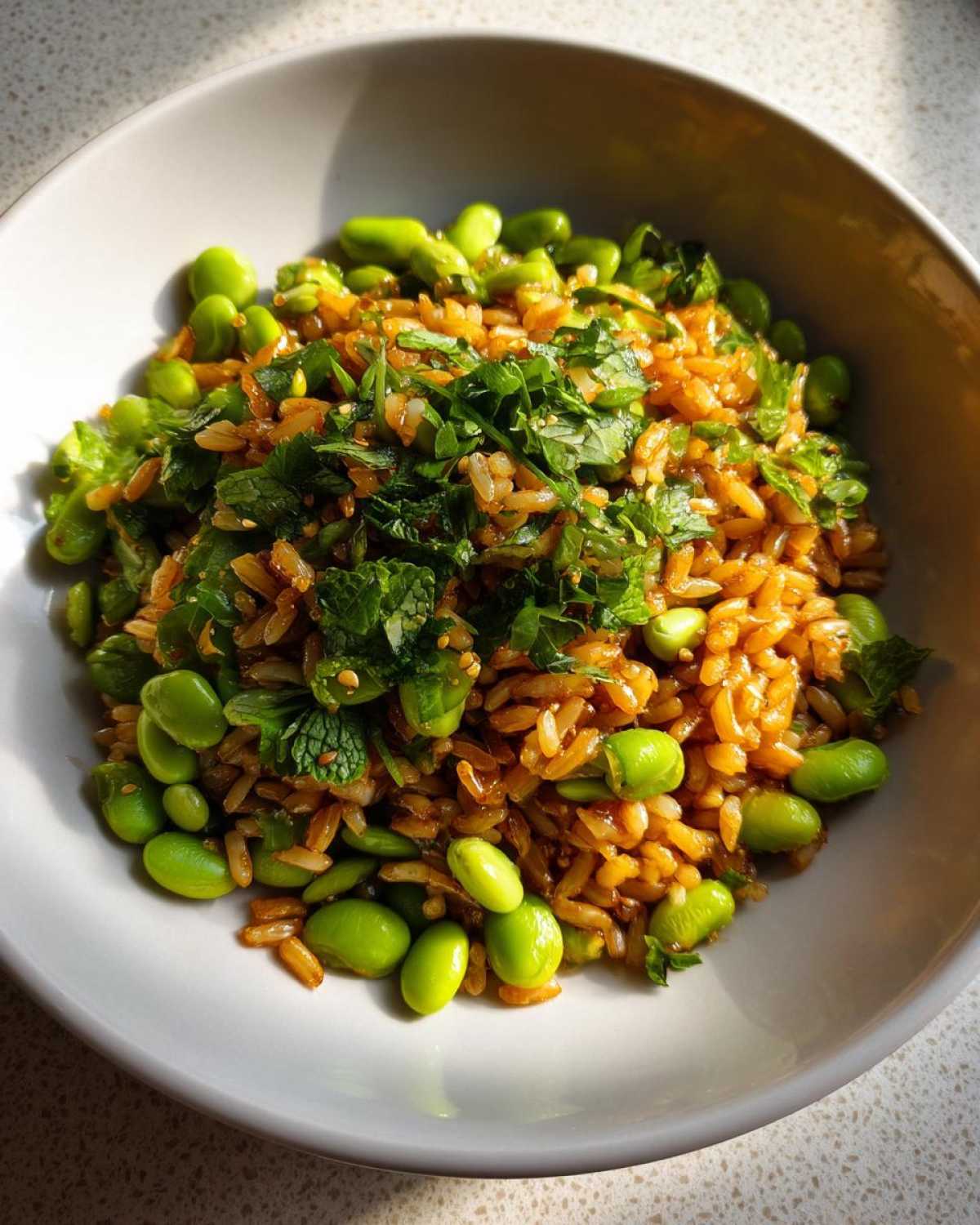 A close-up of a bowl filled with seasoned, slightly browned crispy rice salad mixed with bright green edamame and topped with fresh mint and cilantro.
