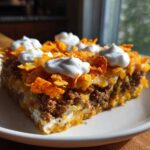 A square slice of Dorito Casserole featuring layers of seasoned ground beef, cheese, and topped with crushed chips and dollops of sour cream.