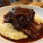 A close-up of tender braised short ribs smothered in rich, dark sauce, served atop a bed of creamy polenta in a white bowl.