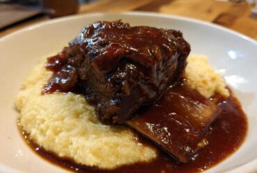 A close-up of tender braised short ribs smothered in rich, dark sauce, served atop a bed of creamy polenta in a white bowl.