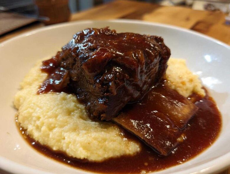 A close-up of tender braised short ribs smothered in rich, dark sauce, served atop a bed of creamy polenta in a white bowl.
