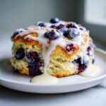 A close-up of a flaky blueberry scone topped with fresh blueberries and a thick vanilla glaze drizzle.