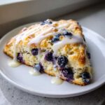 A close-up of a flaky blueberry scone topped with a generous vanilla glaze, bursting with fresh blueberries.