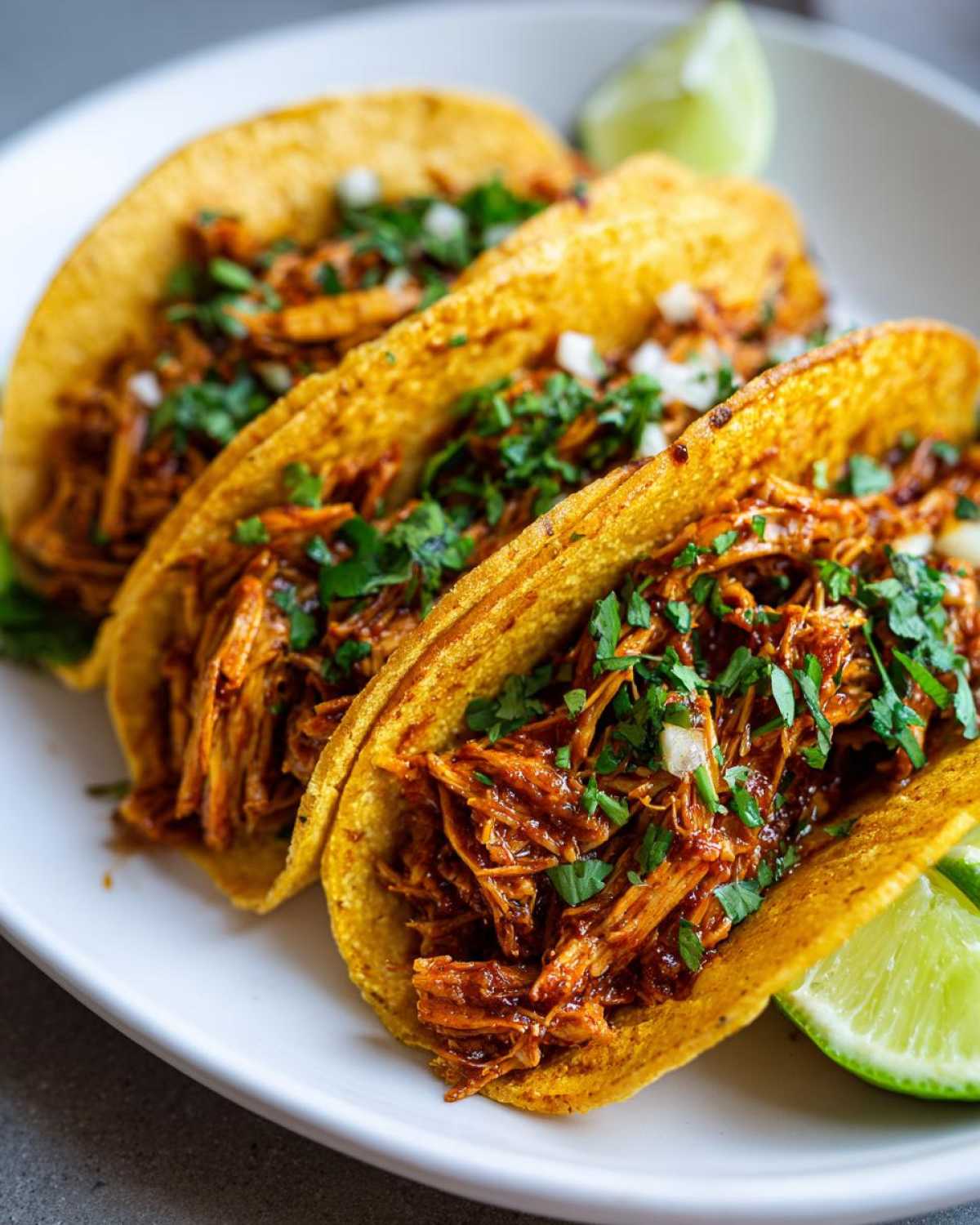 Close-up of three crispy corn shell chicken tacos filled with shredded seasoned chicken, topped with fresh cilantro and onion, served with lime wedges.