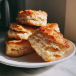 A stack of golden brown, flaky buttermilk biscuits, with one split open showing the soft interior.