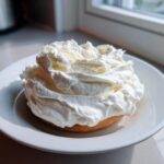 A thick swirl of bright white cream cheese frosting generously topping a small baked good on a white plate.