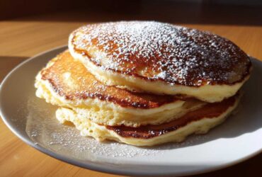 A stack of three fluffy greek yogurt pancakes dusted generously with powdered sugar, sitting on a white plate.