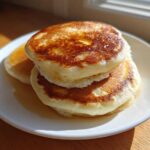 A stack of three thick, fluffy pancakes drizzled with syrup, sitting on a white plate.