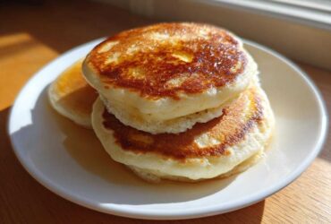 A stack of three thick, fluffy pancakes drizzled with syrup, sitting on a white plate.