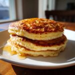 A close-up shot of three fluffy pancakes stacked on a white plate with syrup dripping down the side, highlighting their golden brown tops.