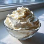 A close-up of light, fluffy vanilla buttercream frosting piped high in a clear glass bowl, backlit by sunlight.