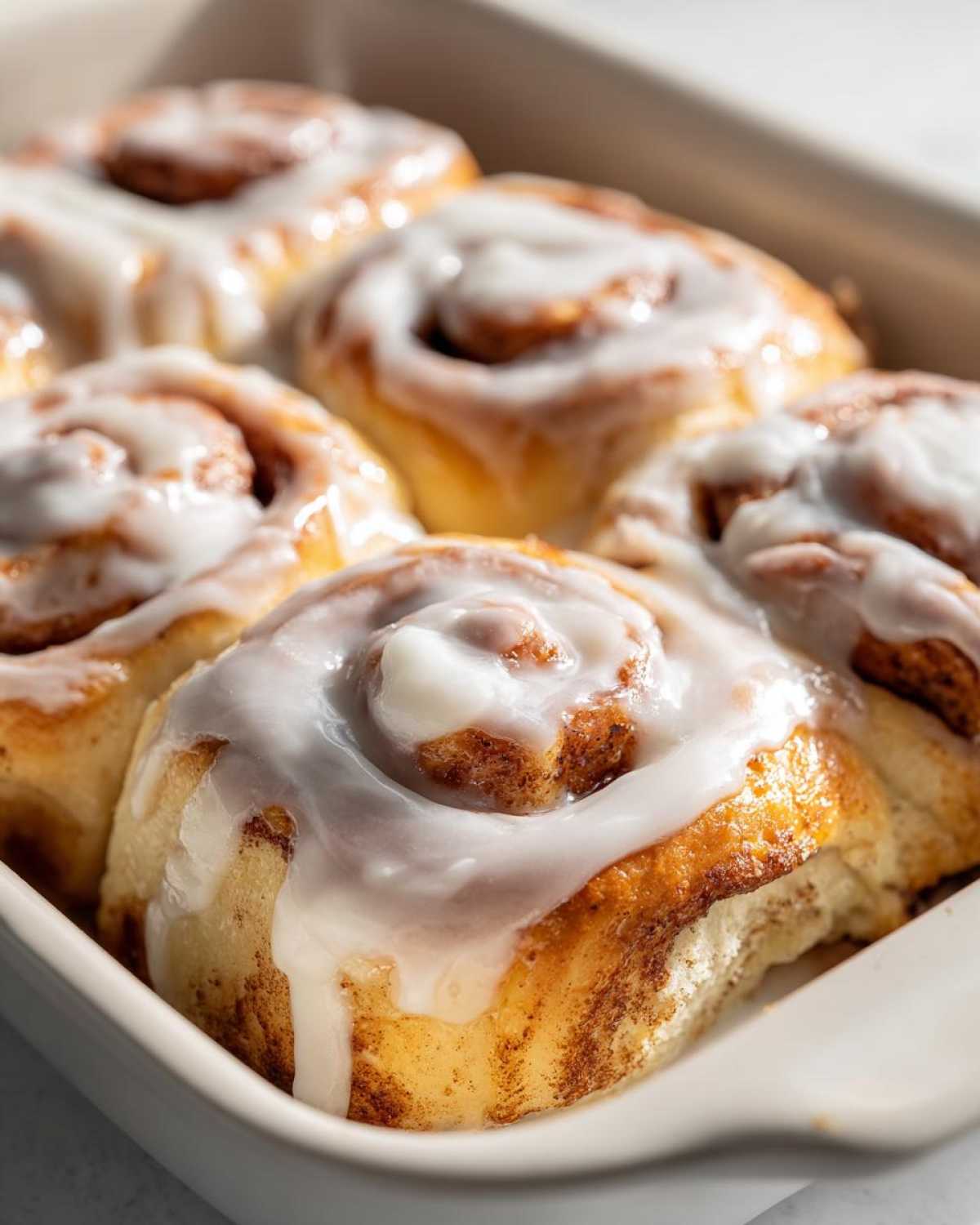 Close-up of several gooey, freshly baked cinnamon rolls topped with thick white icing in a white baking dish.