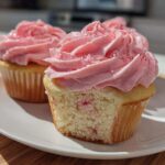 Close-up of fresh strawberry cupcakes, one with a bite taken out showing the cake texture and pink frosting.