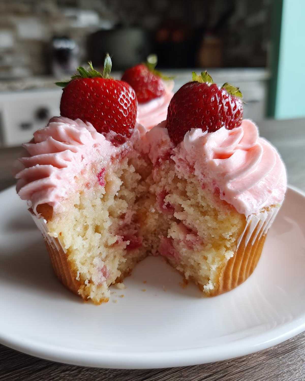 A fresh strawberry cupcake cut in half showing the moist interior with strawberry pieces, topped with pink frosting and a whole strawberry.