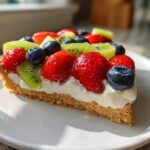 A close-up shot of a slice of fruit pizza sugar cookie topped with cream cheese frosting, strawberries, blueberries, and kiwi.