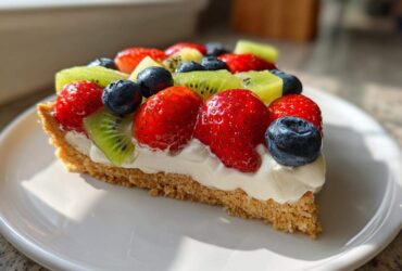 A close-up shot of a slice of fruit pizza sugar cookie topped with cream cheese frosting, strawberries, blueberries, and kiwi.