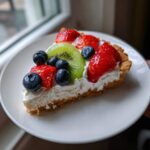 A slice of fruit pizza sugar cookie topped with cream cheese frosting, strawberries, blueberries, and kiwi.
